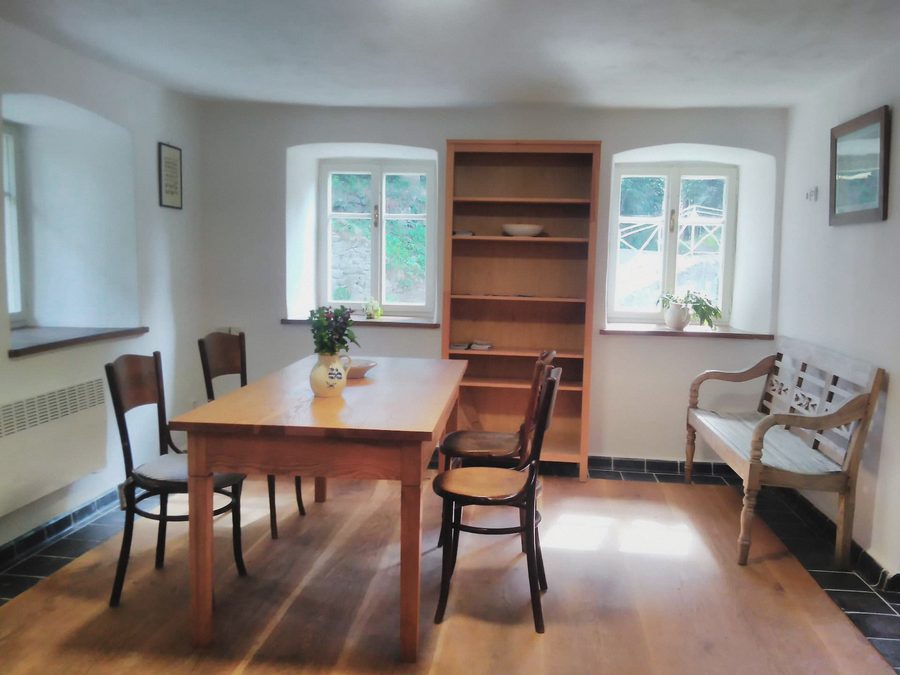 Světlá světnice s dřevěným stolem, židlemi, knihovnou a lavicí